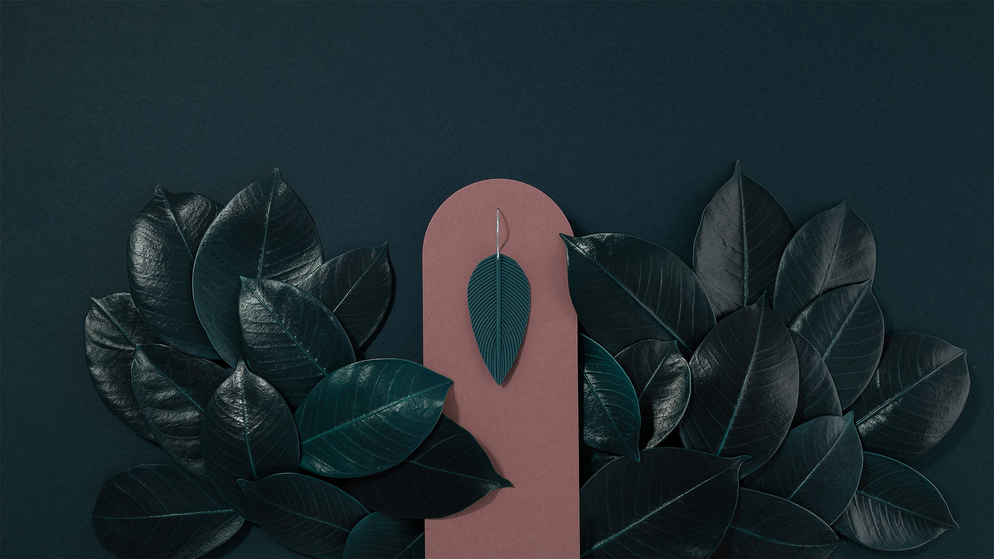 Folium N°1 handmade dangle earring from Portamira’s Folium collection – smokey teal leaf shape with 925 sterling silver hook, on a dark pink display and dark teal leafy background.
