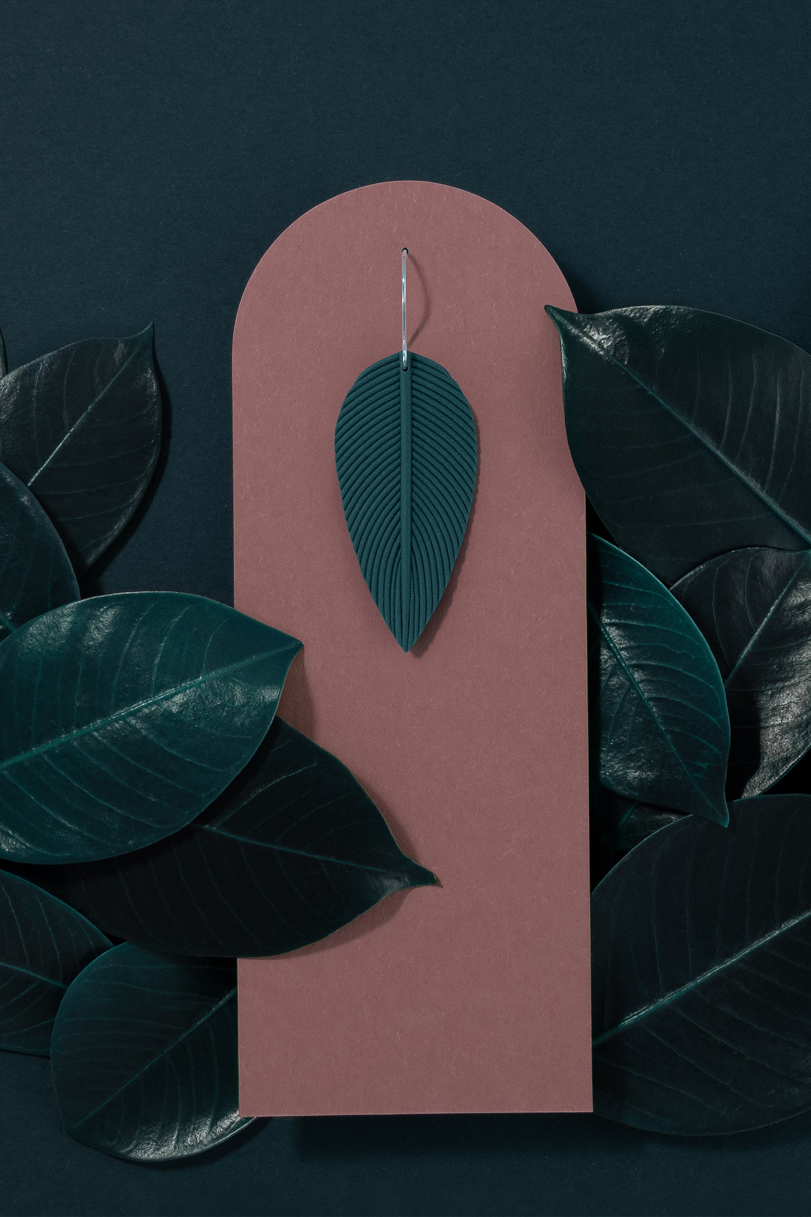 Folium N°1 handmade dangle earring from Portamira’s Folium collection – smokey teal leaf shape with 925 sterling silver hook, on a dark pink display and dark teal leafy background.
