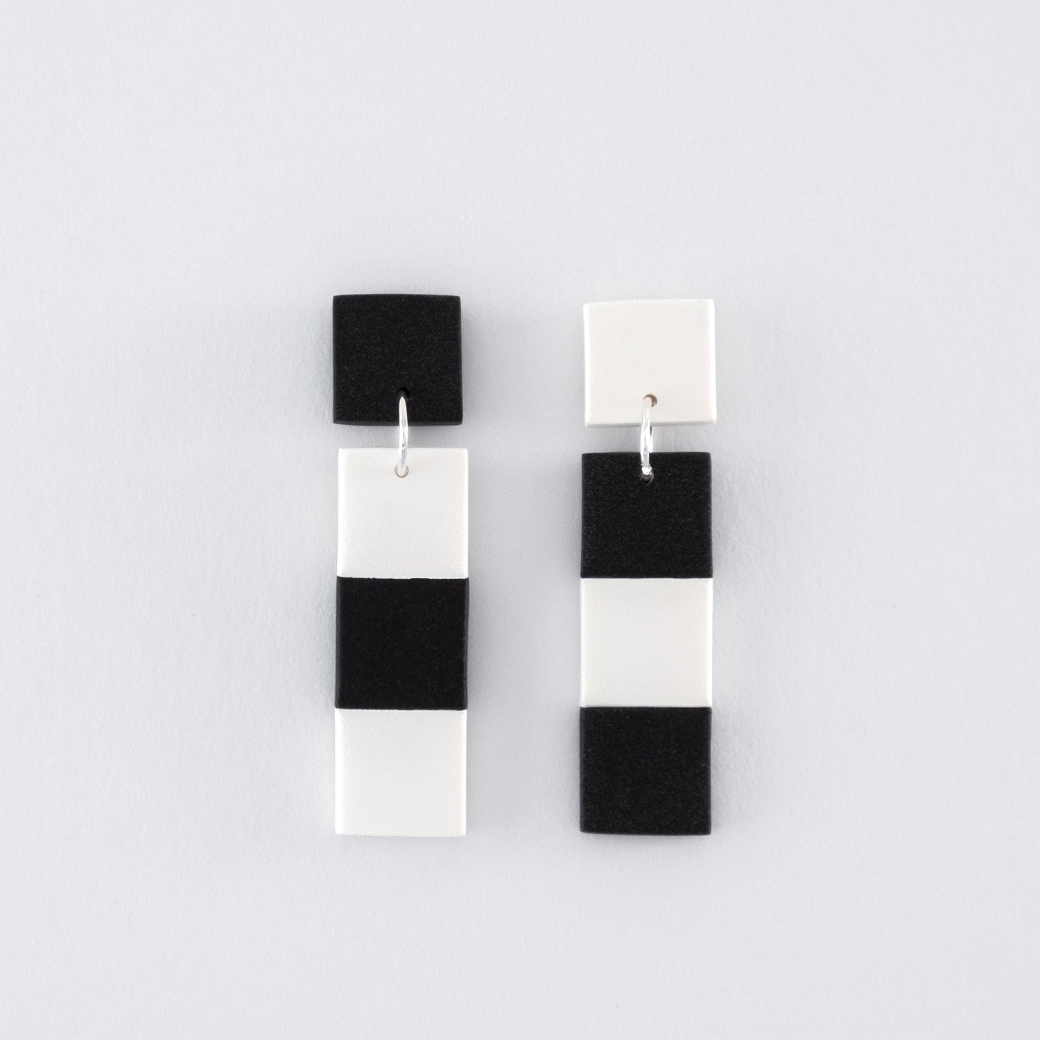A mismatched pair of Black & White N°2 handmade dangle earrings – elongated geometric shapes with black and white checks and 925 sterling silver findings on a light grey background.